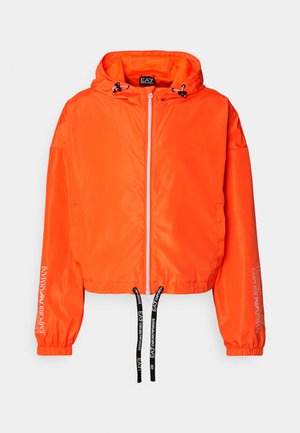 Lyst orange kort jakke med hætte, lavet af glat stof. Har sorte snore og hvid logo tekst på ærmerne og lynlåsen.