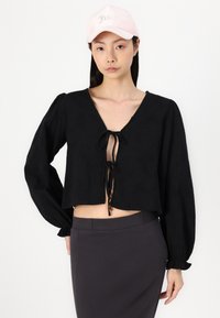 Blusa nera corta con maniche lunghe e voluminose e chiusura a fiocco sul collo. Indossata con una gonna grigia scura e un berretto rosa chiaro.