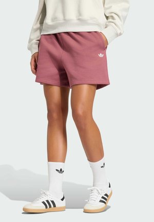 Person iført lilla Adidas-shorts, hvide Adidas-strømper, hvide sneakers med sorte striber og en cremefarvet sweatshirt, stående med den ene hånd i lommen.