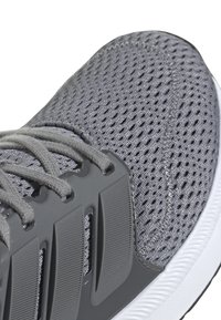 Zapatilla atlética gris con una parte superior de malla, sobrecapas sintéticas texturizadas y cordones grises. Cuenta con una suela de goma blanca con surcos de tracción.