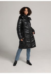 Zwarte pufferjas met hoge kraag en ritssluiting aan de voorkant, voorzien van diagonale naden en zijzakken, gecombineerd met lichtblauwe jeans en enkellaarsjes.