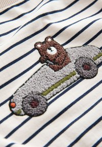 Orso bruno ricamato che guida un’auto grigia su tessuto bianco con sottili righe orizzontali blu navy.