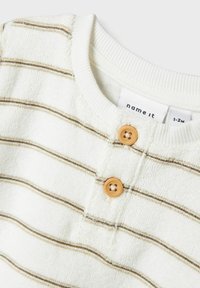 Chemise en coton blanche avec de fines rayures marron, dotée d'un col côtelé et de deux boutons en bois sur le devant. Tissu doux et texturé.