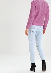 Roze gebreide trui met een textuurpatroon, gedragen met lichtblauwe jeans en zwarte hoge laarzen met hakken. Zicht van achteren.