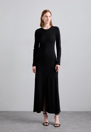 Black long-sleeve dress with a fitted bodice and floor-length skirt. Features a center slit and smooth, stretchy fabric. Black strappy sandals.