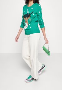 Pull vert en tricot avec un motif de rennes et des pois blancs, associé à un pantalon large crème et des baskets multicolores, tenant un sac à main vert.
