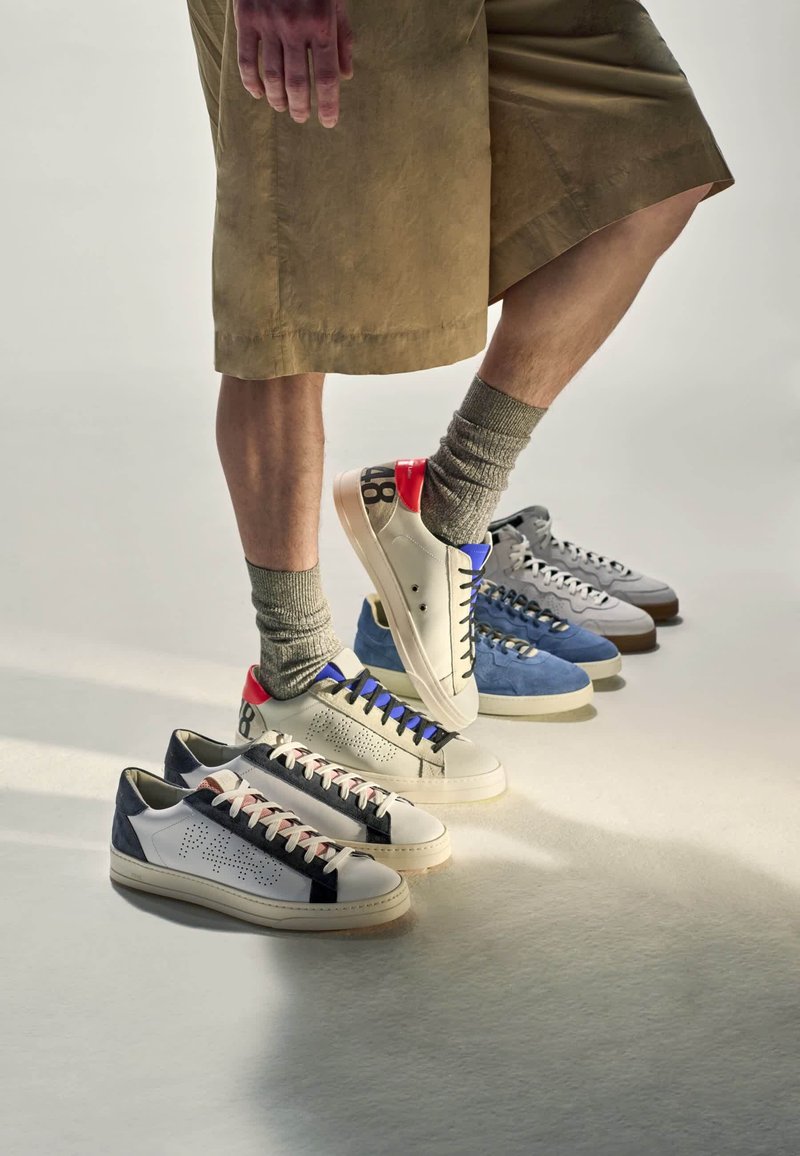 Personne portant un short beige et des chaussettes grises marche sur des baskets blanches avec des détails colorés alignées avec d’autres paires grises et bleues sur un sol pâle.