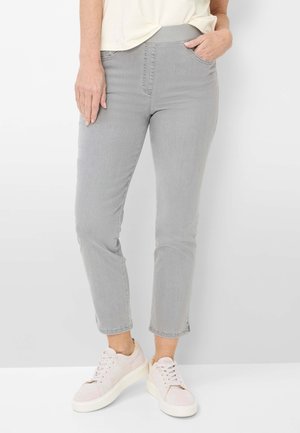 Kvinde iført lysegrå jeans til anklerne med elastisk talje og lyserøde sneakers, hånd i lommen, står op ad en hvid væg.