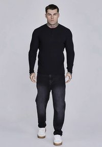 Zwart langärmelig henley-shirt met een knoopsluiting bij de hals, gecombineerd met een donkere spijkerbroek en witte sneakers, heeft een aansluitend ontwerp en een geribbelde textuur.