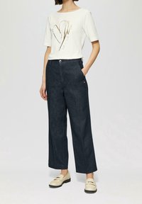 Donna che indossa una maglietta bianca a maniche corte con un design a cuore dorato, pantaloni larghi scuri e mocassini crema, in piedi con una mano nella tasca.