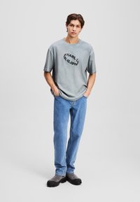 T-shirt en coton gris avec un logo noir rond, associé à un jean en denim bleu clair et des bottes grises, sur un fond neutre.