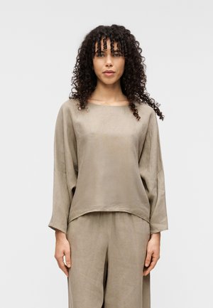 Bluză lejeră bej din in, cu mâneci lungi, decolteu rotund și tiv ușor scurtat, asortată cu pantaloni din același material de in. Textură fină.