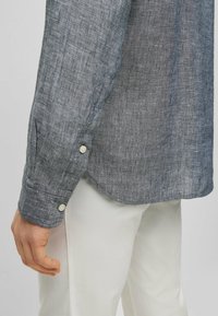 Grå linneskjorta med en texturerad yta, med knäppte manschetter och böjd fåll, i kombination med vita byxor.