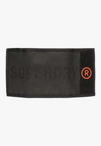 Czarna tkaninowa łatka z wytłoczonym napisem "SUPERDRY" w macie, z sekcją na rzep oraz małym czerwonym okrągłym logo po prawej stronie.