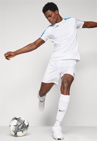Hvit fotballdrakt med blå detaljer, inkludert en t-skjorte med korte ermer og shorts. Spiller i bevegelse med en svart og hvit fotball.