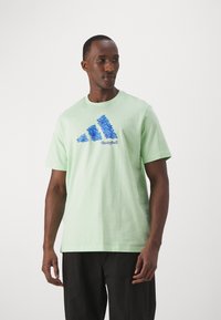 T-shirt in cotone verde chiaro con una grafica floreale blu di tre strisce e la parola "Basketball" stampata sotto. Maniche corte, collo girocollo.