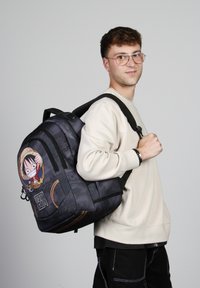 Mochila de tela negra con diseño de personaje, que presenta una figura de dibujos animados en la parte frontal. Correas ajustables y detalles en la superficie texturizada.