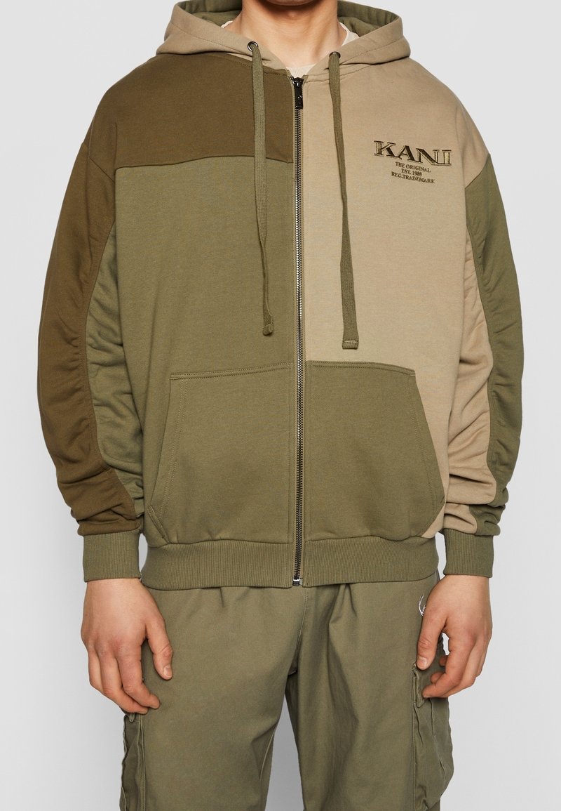 Homme portant un sweat à capuche zippé vert multitone et beige avec des poches devant et le texte "KAN II" sur la poitrine.