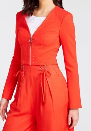 Rode cropped jas met lange mouwen, een ritssluiting aan de voorkant en zilveren studs langs de halslijn. Gecombineerd met losse broek met veters als detail.