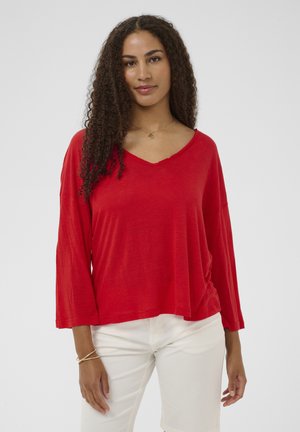 Žena s kučeravými vlasmi, oblečená v voľnom červenom tričku s výstrihom do V a bielych nohaviciach, stojaca pred jednoduchým svetlým pozadím.