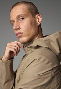 Beige wasserdichte Jacke mit Kapuze, ausgestattet mit einem hohen Kragen und sichtbaren Nähdetails. Modell, das den Kragen leicht mit einer Hand festhält.