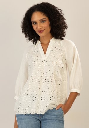 Woman with curly hair wearing a white embroidered blouse with 3/4 sleeves and blue jeans, posing against a plain background.