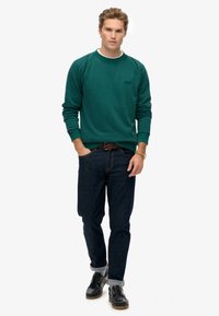Groene sweatshirt met ronde halslijn en geribbelde manchetten, gecombineerd met donkerblauwe spijkerbroek. Model draagt zwarte schoenen met veters en opgerolde manchetten.