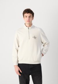 Pullover crema chiaro con colletto a mezza zip, polsini e orlo a coste, con un dettaglio del logo scuro sul petto e una texture liscia.