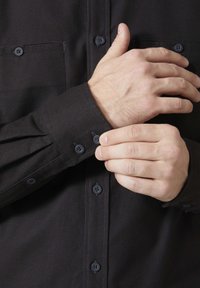 Zwarte overdingenhemd met lange mouwen en twee knopen bij de manchetten. De stof lijkt glad, met subtiele stiksels op de knoopsluiting.