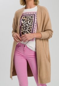 Cardigan beige a maglia con apertura frontale e maniche aderenti. Sotto, una maglietta bianca con un motivo leopardato in nero e rosa. Pantaloni rosa.