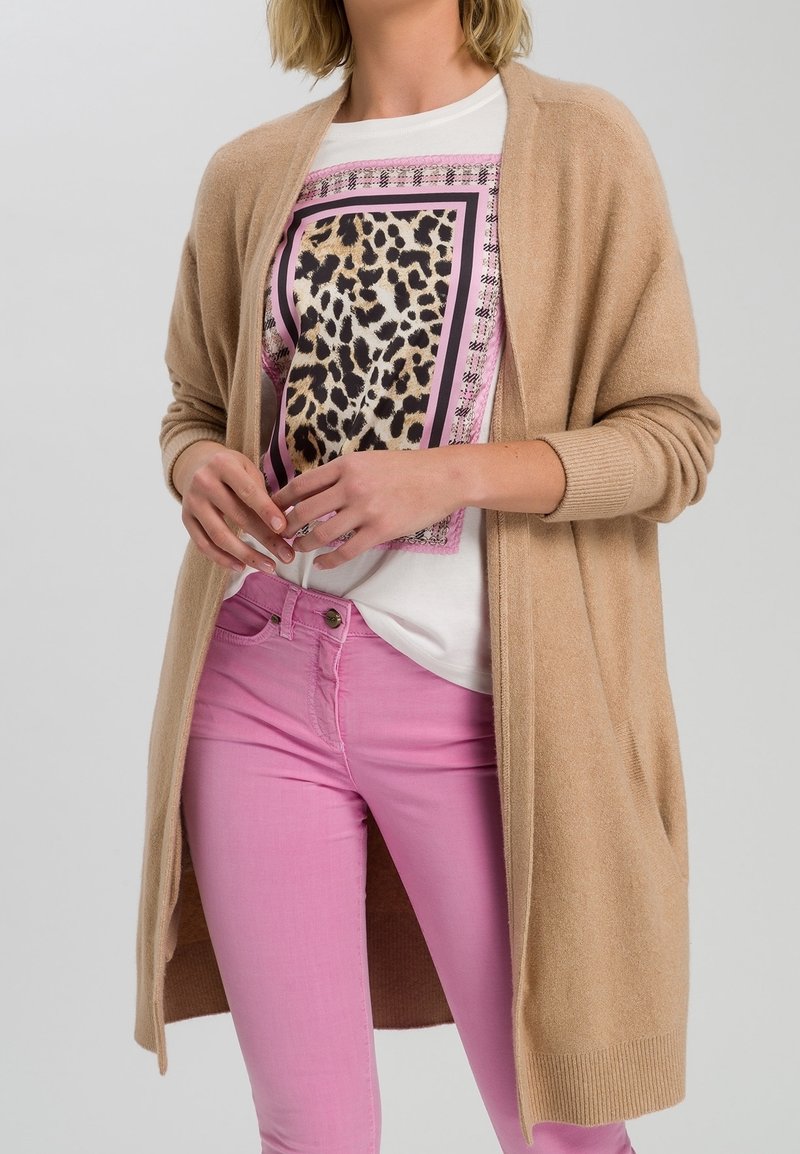 Cardigan beige a maglia con apertura frontale e maniche aderenti. Sotto, una maglietta bianca con un motivo leopardato in nero e rosa. Pantaloni rosa.