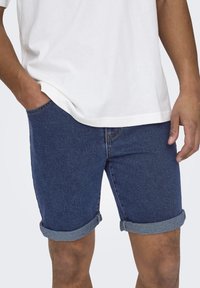 Shorts di jeans blu scuro con orlo arrotolato, caratterizzati da cinque tasche e cuciture a contrasto. Indossati con una t-shirt bianca semplice.