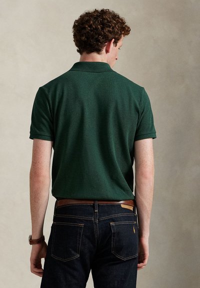 Homme aux cheveux bouclés portant un polo vert foncé à manches courtes rentré dans un jean bleu foncé avec une ceinture marron, de dos.