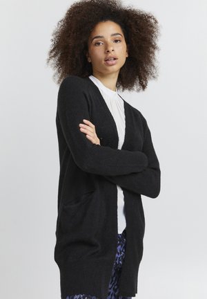 Cardigan lungo nero realizzato in morbido tessuto a maglia, con due tasche anteriori e una vestibilità rilassata, indossato sopra una camicia bianca con colletto.