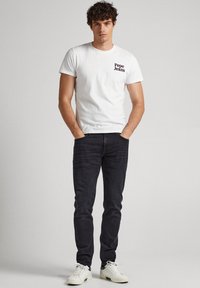 Jongeman staat met zijn handen in zijn zakken, draagt een wit Pepe Jeans T-shirt, zwarte jeans en witte sneakers voor een effen achtergrond.