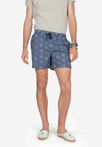 Bañadores azules con un patrón de pulpos y calaveras, con cintura ajustable y acentos en color sólido. Combinados con zapatos de lona ligeros.