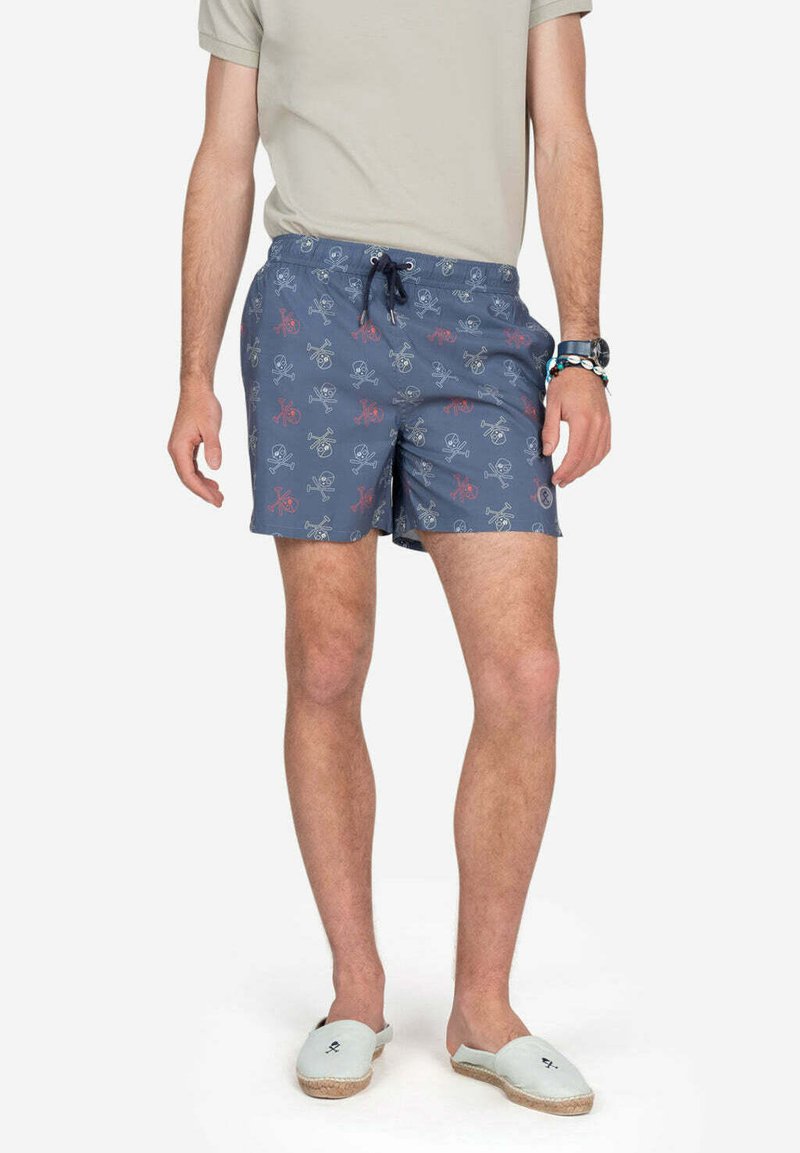 Bañadores azules con un patrón de pulpos y calaveras, con cintura ajustable y acentos en color sólido. Combinados con zapatos de lona ligeros.