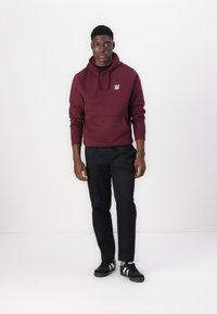 Sweat à capuche bordeaux avec une poche devant, arborant un petit logo. Associé à un pantalon noir et des baskets noires avec des rayures blanches.
