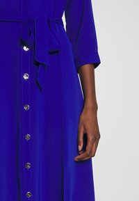 Blå klänning med bälte i midjan, med knapplås framtill och en sidoklyfta. Tyget verkar slätt med en lätt glans. Långa ärmar.