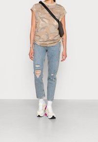 Camisa beige con estampado de camuflaje y manga corta, combinada con unos vaqueros desgastados de color azul claro que tienen detalles rasgados. Terminada con zapatillas blancas.