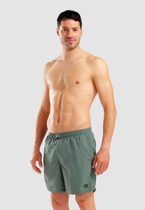 Hemdloser Mann mit kurzen dunklen Haaren, der grüne Badeshorts trägt und vor schlichtem grauem Hintergrund steht.