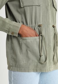 Olive green jacket featuring two chest pockets, drawstring waist, and button closures on sleeves. Soft, textured fabric. Casual design.