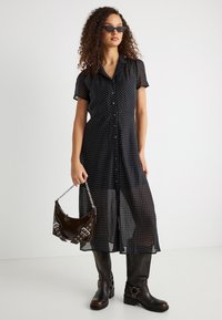 Γυναίκα που φορά μαύρο διαφανές μίντι φόρεμα με πουά, μαύρες μπότες μέχρι το γόνατο, μαύρα γυαλιά ηλίου και κρατά μικρή μαύρη τσάντα με ασημί αλυσίδα.