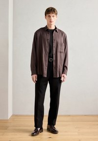 Camisa de botones a rayas marrones con un bolsillo en el pecho, combinada con pantalones negros y zapatos oscuros. Textura suave, corte clásico, estilo casual.