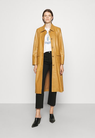 Long mustard leather coat with button closure, side pockets, and a straight cut. Worn over a graphic t-shirt and black cropped trousers.