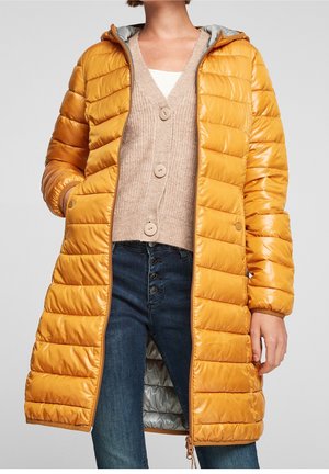 Femme portant une longue doudoune matelassée jaune moutarde par-dessus un cardigan beige boutonné et un jean bleu marine taille haute.