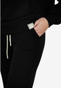 Czarna bluza z kapturem i dresy z boczną kieszenią, wyposażone w białą i szaro-zygzakową metkę. Sznurek w talii z jasnymi końcówkami.