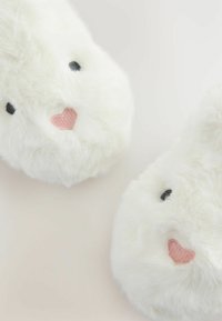 Weiße, flauschige Hausschuhe in Form von Hasengesichtern, mit schwarzen gestickten Augen und pinken Herz-Akzenten auf der Nase. Weicher, plüschiger Stoff.