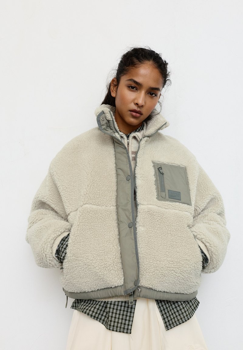 Lysebeige faux shearling jakke med grå, polstret krave, lynlåslomme og elastisk kant. Båret over en ternet skjorte.