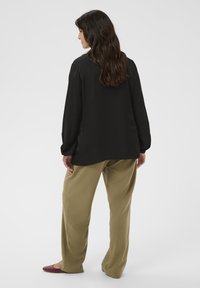 Camicia nera a maniche lunghe con una texture liscia, indossata con pantaloni a gamba larga verde oliva. Include dettagli sottili sulle spalle, girata di spalle rispetto alla fotocamera.